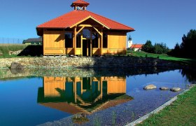 Graduation facility Waldenstein, © Gradieranlage Waldenstein A wooden building with a red roof is reflected in a pond surrounded by a green landscape.