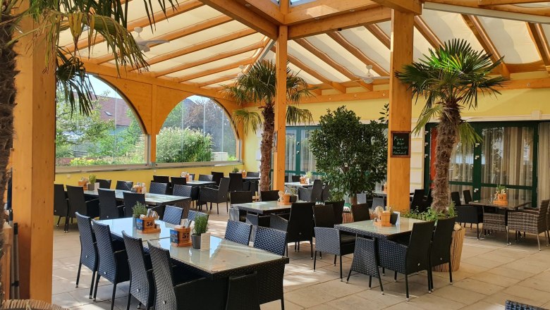 Lifetime Restaurant, © Familie Eissler Covered terrace of a restaurant with palm trees and tables.