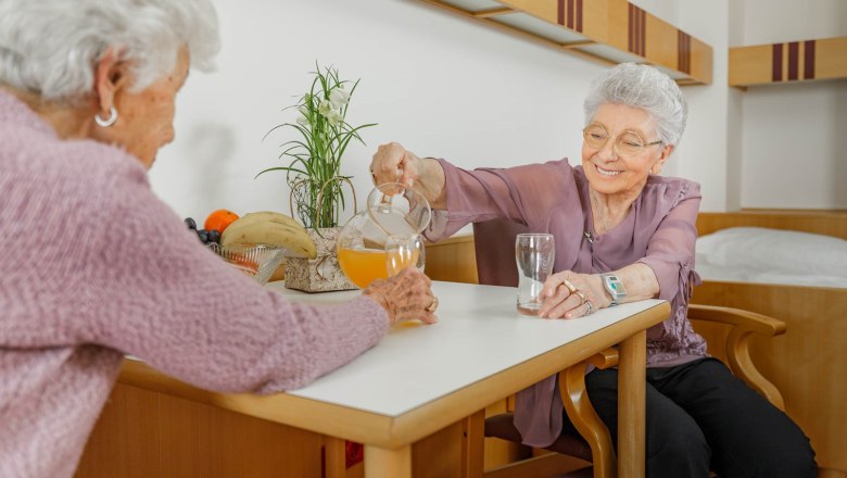 Cozy comfort rooms, © ÖJAB Waldpension - Christian Kremsl Two elderly women are sitting at a table in a cozy room. One woman is pouring juice from a carafe into a glass.