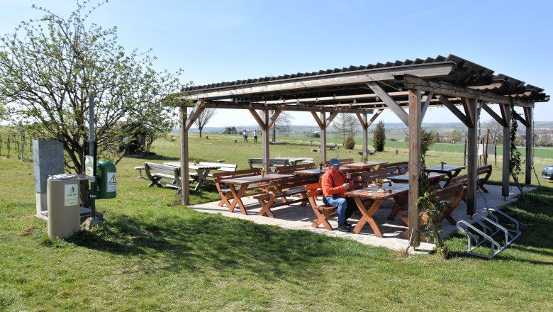 Pleasure rest area at the tree circle, © StadtGemeinde Mistelbach / Mag. Mark Schönmann A covered rest area with wooden benches and tables on a meadow, a person is sitting there. Fields and trees can be seen in the background.