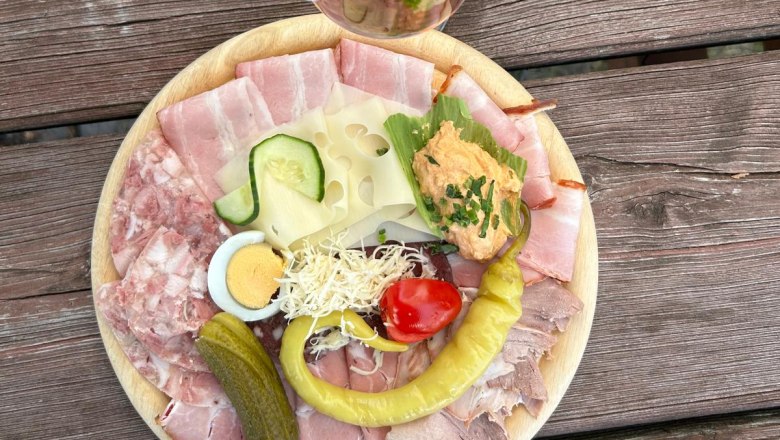 Heurigen enjoyment, © Weingut Döllinger A plate with cold cuts, cheese, egg, cucumber, peppers and spread on a wooden table. Next to it, a glass of rosé wine with a straw.