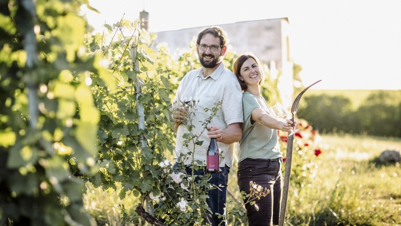 Michael and Monika Sailer, © Astrid Bartl Two people stand smiling in a sunny vineyard with a bottle of wine and a glass.