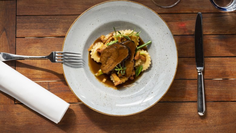 Braised beef cheeks, © Niederösterreich Werbung/Michael Reidinger A plate of braised beef cheeks, ravioli and vegetables on a wooden table, with cutlery and a glass of red wine next to it.