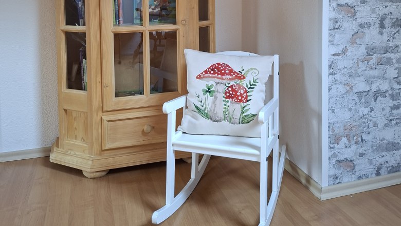 Anteroom, © Manuela Heher A wooden cupboard with books and a white rocking chair with a cushion showing mushrooms stand in a room with a wooden floor and patterned wallpaper.