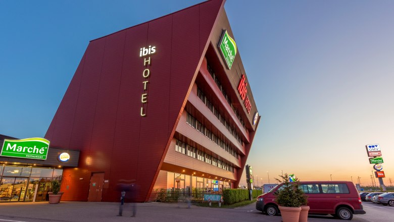 Ibis Vienna Airport, © Marché Restaurants Österreich GmbH Modern hotel building with sloping façade and illuminated lettering 'ibis HOTEL'.