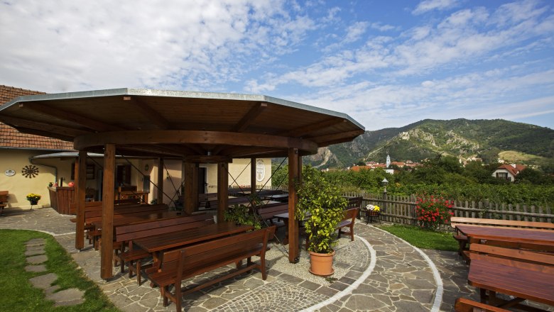 Wine tavern Rehrl-Fischer, © Monika Loeff A guest garden with wooden tables and benches, surrounded by green countryside and mountains in the background.