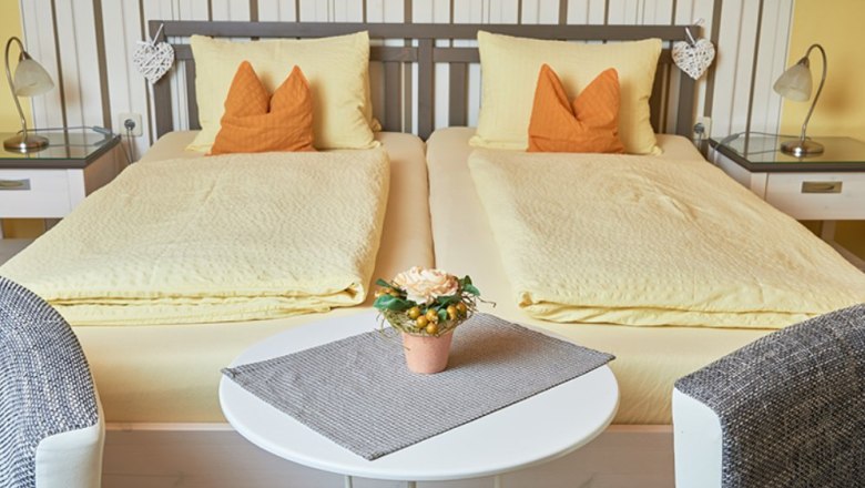Pension Parzer Rooms, © Weinhof Parzer Cozy double room with two single beds, yellow bed linen and orange pillows. A round table with a flower arrangement stands between two armchairs.