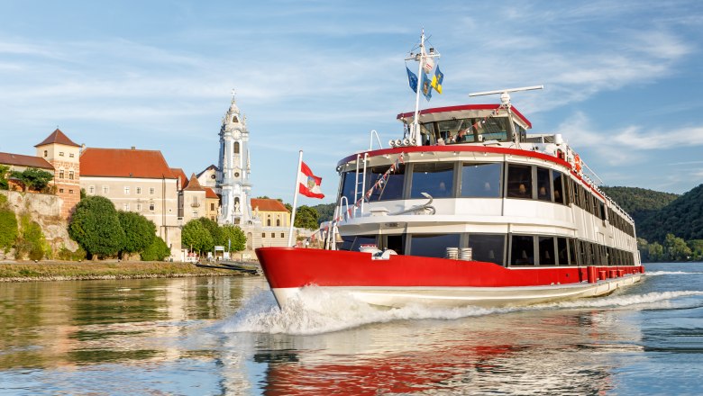 DDSG Blue Danube Shipping, © DDSG Blue Danube Schiffahrt/Wöckinger A red passenger ship sails on the Danube in front of a historic town with a striking church tower.