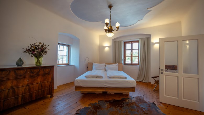 Oberstockstall Estate, © Niederösterreich Werbung / Maximilian Pawlikowsky A cozy bedroom with wooden floor, double bed, antique chest of drawers and bouquet of flowers. Ceiling light and window with curtains.
