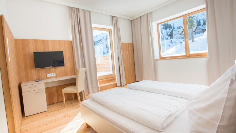 JoSchi Hochkar sports shop, © JoSchi Sporthaus Hochkar Bright hotel room with double bed, desk and view of snow-covered mountains.