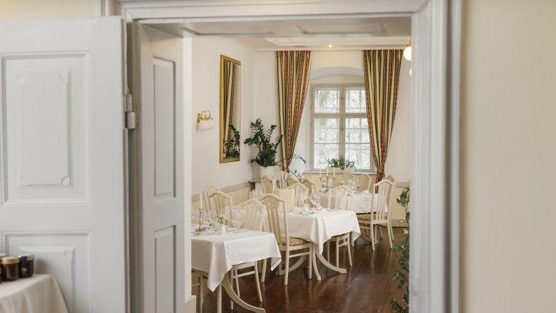 Gloggnitz Castle Restaurant, © Wiener Alpen/ Sophie Menegaldo Elegant restaurant with laid tables, white tablecloths and classic chairs, visible through an open door.