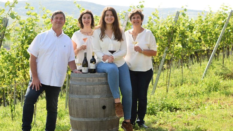 TDR wine tavern Kienberger family, © Kienberger Four people stand and sit in a vineyard, smiling with wine glasses in their hands, next to a wine barrel with bottles on it.