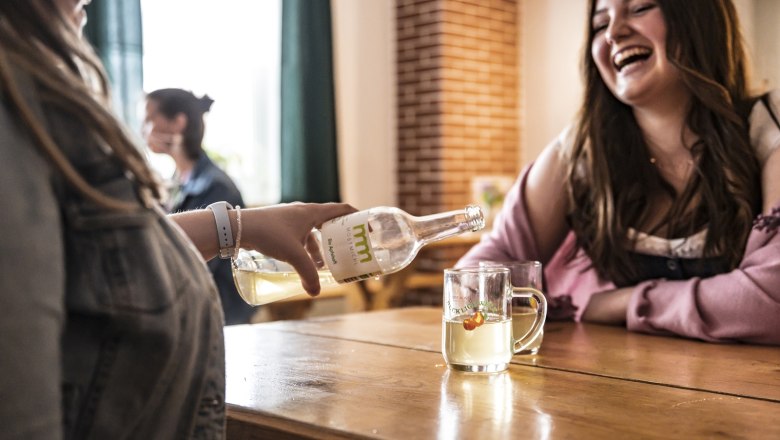 Quality must from our own production, © Thomas Gobauer-Photography Two women are sitting at a table, one is pouring Cider from a bottle into a glass. They are both laughing.