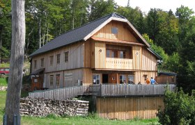 Waldburgangerhütte, © Maria Stroebl A wooden hut in the forest with a terrace and people on it.