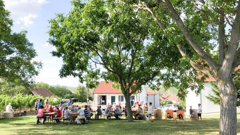 WineDeer, © Wein:Hirsch People sit outside at tables under trees, in the background a building with a red roof.