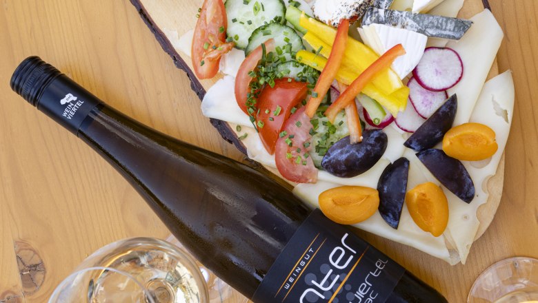 Heurigen platter, © Weinviertel Tourismus GmbH / Schwarz-König A Heurigen platter with various cheeses, spreads, vegetables and a bottle of wine on a wooden table.