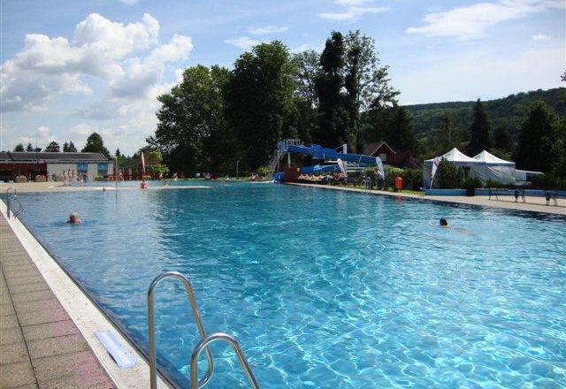 Schlossbad Pitten, © Marktgemeinde Pitten A large outdoor pool with clear water, surrounded by trees and hills under a blue sky.