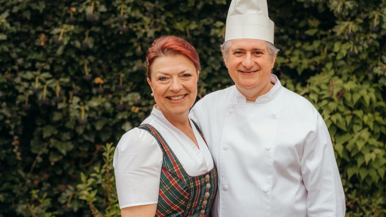 Elvira Hubinger and Oswald Topf, © Niederösterreich Werbung/Daniela Führer A man in chef's clothes and a woman in traditional costume smile at the camera against a backdrop of green leaves.
