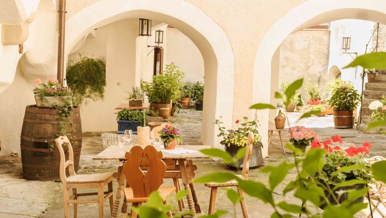 Kirchenwirt Weißenkirchen, © Rita Newman A cozy courtyard with a wooden table and chairs, surrounded by plants and flowers in pots.