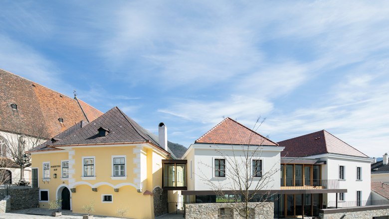 Mauritiushof wine experience world, © Weingut FJ Gritsch Two buildings with yellow and white facades, connected by a modern glass corridor, under a blue sky.