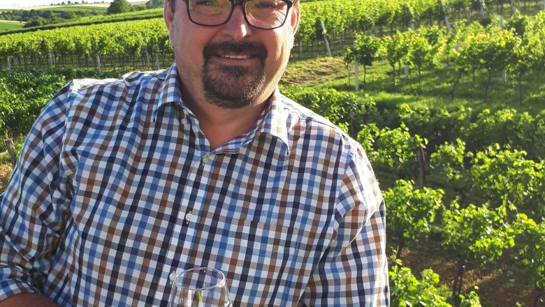 winemaker-family-resch, © Familie Resch A man in a checked shirt stands in front of a vineyard with a wine glass in his hand.