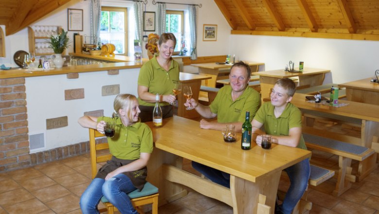 Everyone pitches in at the wine tavern, © Astrid Bartl A family in green shirts sits at a wooden table in a wine tavern and toasts with drinks.
