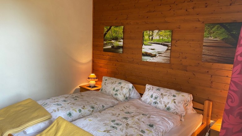 Double room, © Wiener Alpen Cozy double room with wooden walls, a double bed with floral bedding and yellow blankets, as well as a bedside lamp.