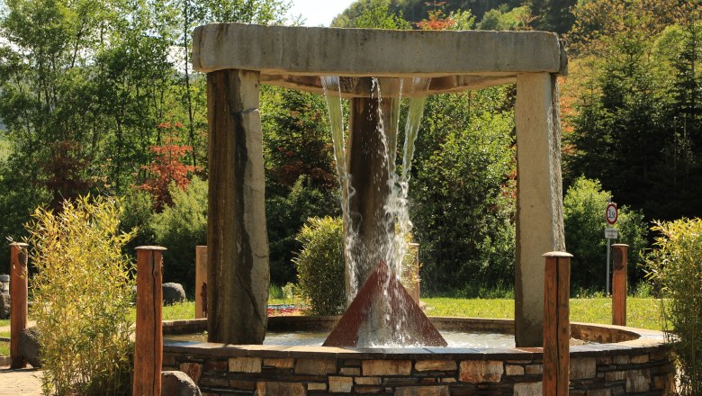 Fountain in the Celtic Park, © Marktgemeinde Schwarzenbach, Helmut Karner A modern outdoor fountain with a pyramid-shaped structure in the center, surrounded by trees and plants.
