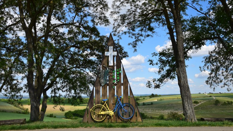 Cyclist Marterl in Mittergrabern, © Christian Häusler A shrine with a yellow and blue bicycle, surrounded by trees and fields under a blue sky.