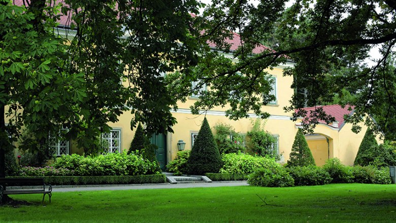Park, © Gärtner Starkl A yellow building with red roofs, surrounded by green trees and manicured lawns in a park.