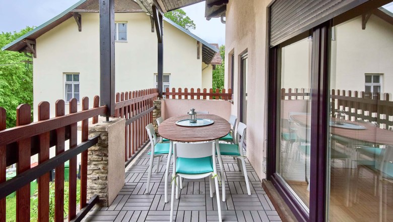 Top 2 - Balcony, © VP FeWo OG A balcony with a wooden table and chairs, surrounded by a wooden fence, with a view of a neighboring building.