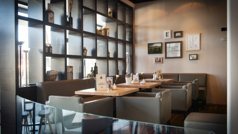 Café Haubiversum, © Haubis GmbH Interior view of a modern café with wooden tables and gray chairs, decorated with plants and pictures on the wall.