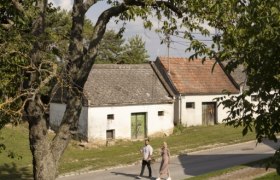 Wine cellar lane in Kettlasbrunn, © Weinviertel Tourismus / Schwarz-König Two people walk past old buildings in a rural setting.
