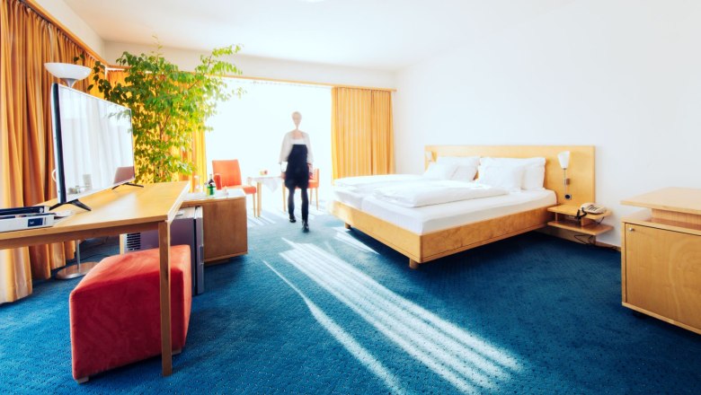 Living space_3, © Klinglhuber Bright hotel room with double bed, desk, TV and large plant. One person walks through the room.