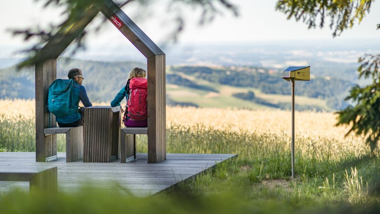 Rest area near the lookout point at Hutwisch, © Wiener Alpen/Martin Fülöp Two hikers sit on a wooden platform with a view of a hilly landscape.