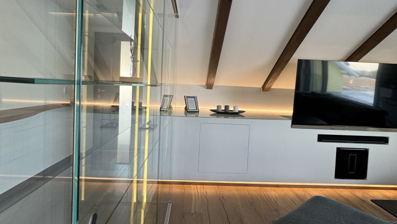Spacious living room, © Juwel am Stadtrand Modern living room with wooden floor, glass cabinet, TV and sloping ceiling with wooden beams.