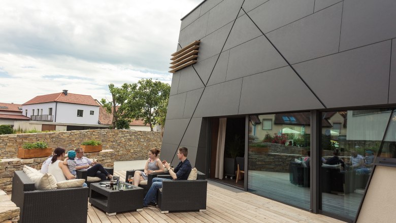 Wooden terrace of the wine lodge, © KASTNER NEW MEDIA - kastner.tv People sitting on a terrace in front of a modern building.