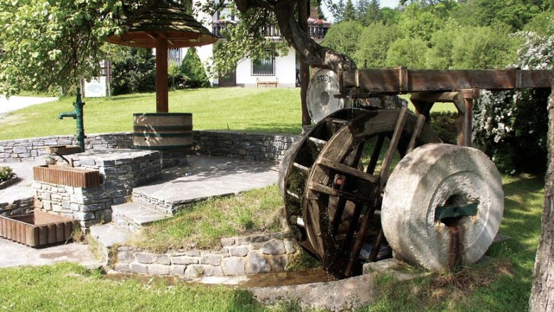 Mill wheel, © Gasthaus Buchegger, Foto Buchegger Old mill wheel with millstone, next to steps and a fountain, surrounded by meadow and trees.