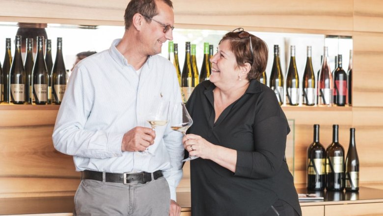 Wolfgang and Elfi Aigner, © Michael Parak A man and a woman toast with wine glasses, wine bottles in the background.