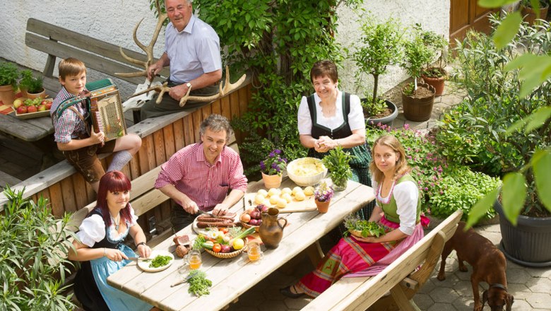 Get together for a snack in the guest garden, © © Langthaler A family in traditional dress sits at a table in the garden and prepares a meal.