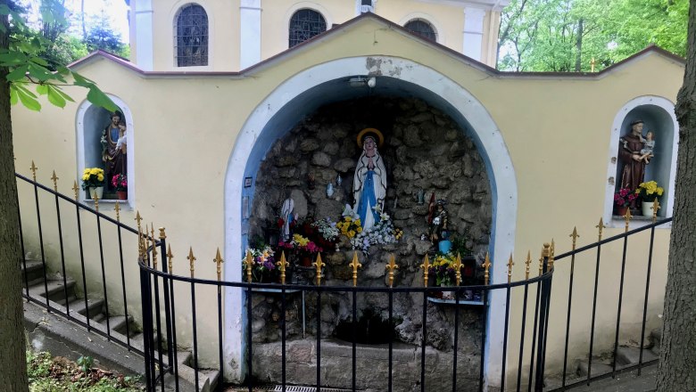 The water in the Bründl Grotto is said to have healing powers, © Weinstraße Weinviertel A grotto with religious statues and flowers, surrounded by a wrought-iron fence.