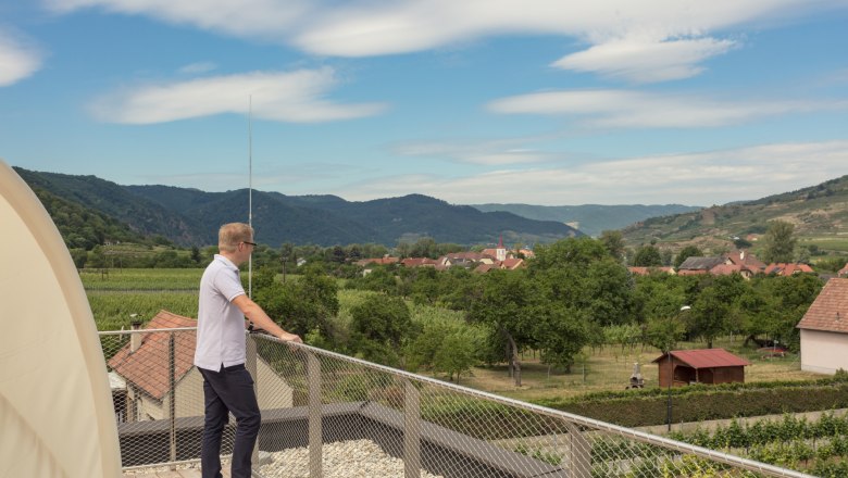 View of the vineyards from the terrace, © KASTNER NEW MEDIA - kastner.tv Man standing on terrace with view of village and hills.