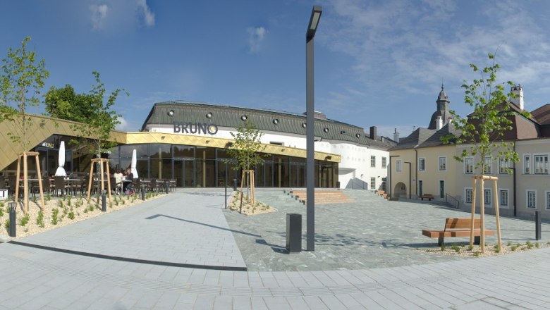 BRUNO, © Prinz Wolfgang Photography Modern architecture with golden facade and traditional building in the background, blue sky.