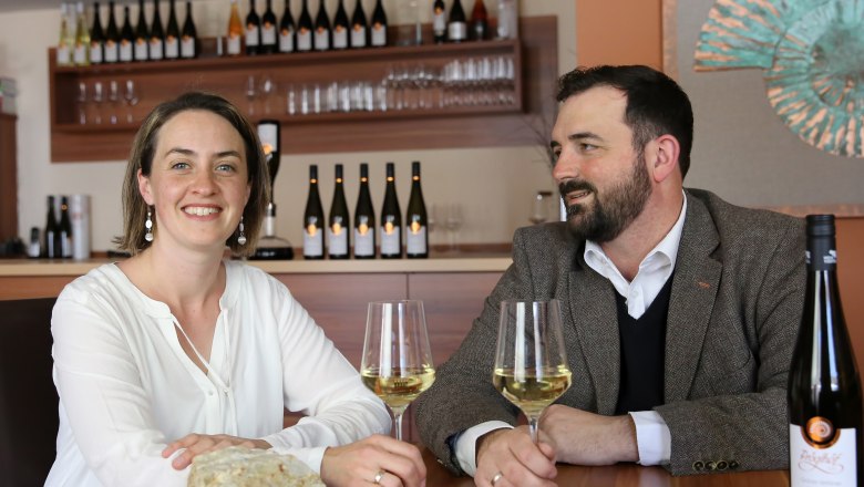 Marianne and Markus Pröglhöf, © Matthias Karasek Two people are sitting at a table with wine glasses and bottles of wine in the background.
