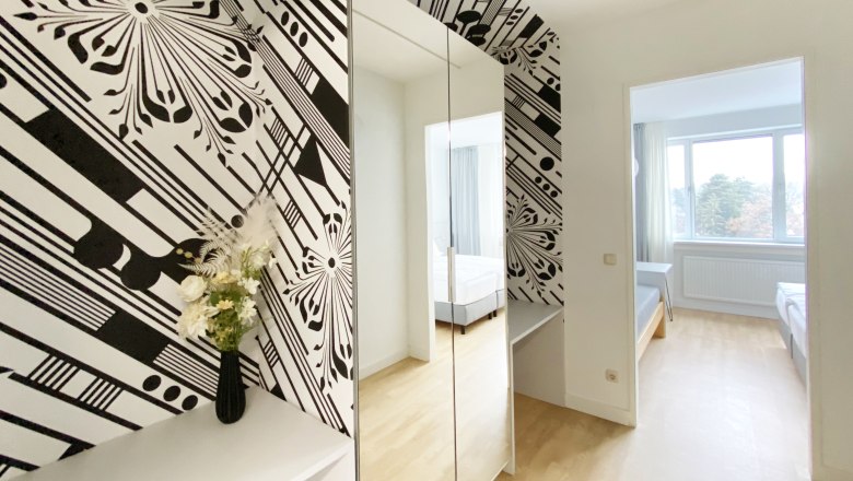 Guest room, © Stadthotel Hollabrunn Modern guest room with black and white wallpaper, mirror, flowers and a view of a bed through a door.