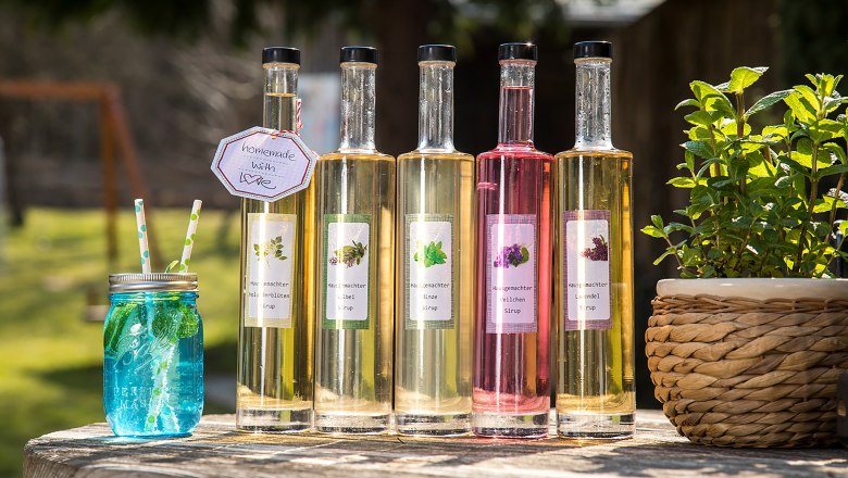 Homemade syrups from the herb garden of Gasthaus Rath-Smetana, © Niederösterreich Werbung/Kurt Pinter Five bottles of homemade syrup and a blue glass with straws on a wooden table outside.