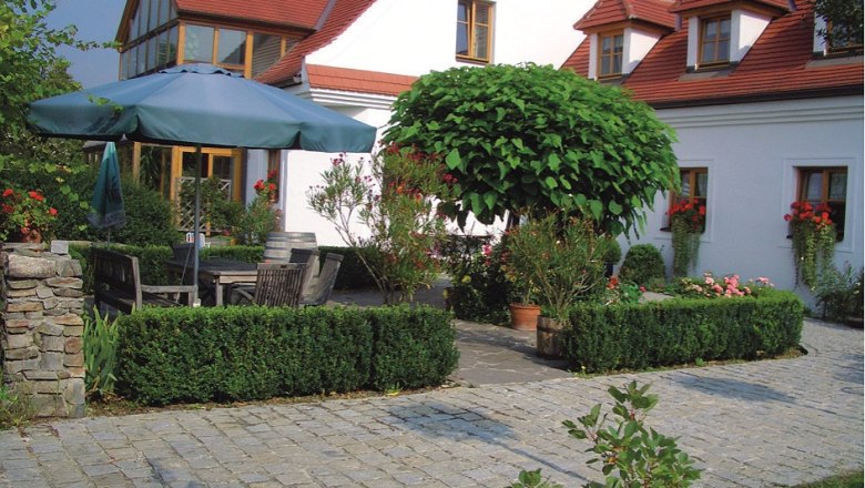 Prabatsch-Aichinger winery, © Winzergut Prabatsch-Aichinger A well-kept garden with paving stones, a large parasol, wooden furniture and a white building with red roofs in the background.