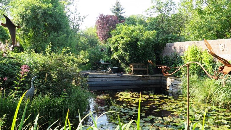 Place by the pond, © Natur im Garten/Martina Liehl-Rainer An idyllic garden with a pond, water lilies and lush vegetation. In the background are a table and chairs on a terrace.