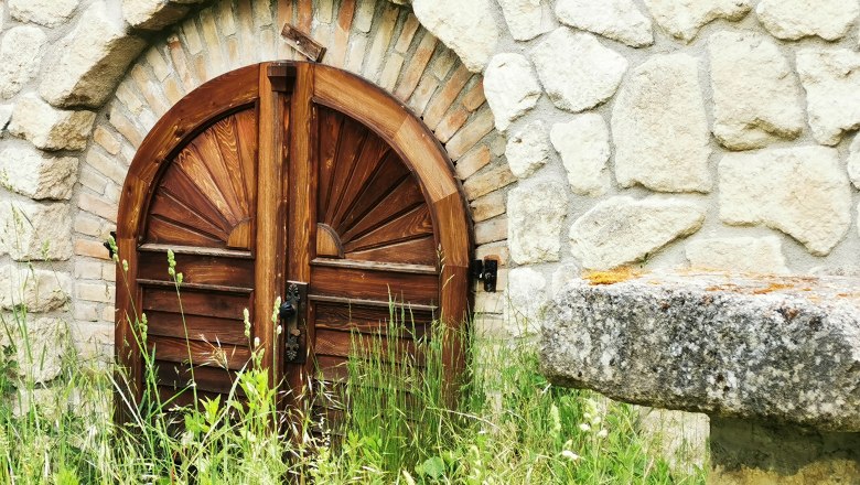 Lovingly restored and hidden behind tall grass is a cellar, © Weinstraße Weinviertel Wooden door in stone wall with grass in the foreground.