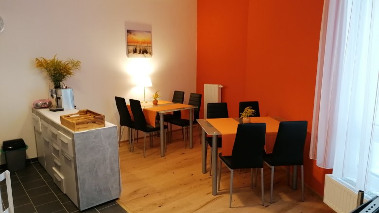 Breakfast room, © Pension zum Spitz A breakfast room with two tables, black chairs and orange tablecloths. A chest of drawers with a coffee machine is on the left.